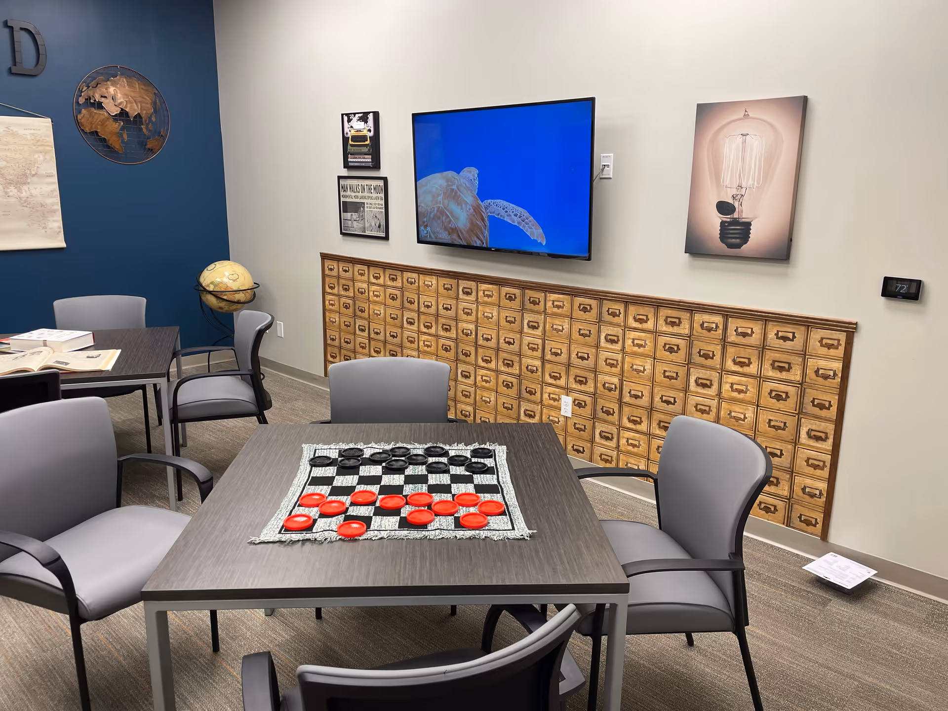 Communal activity room with tables, chairs, a checkerboard, globe decor, and a wall-mounted TV