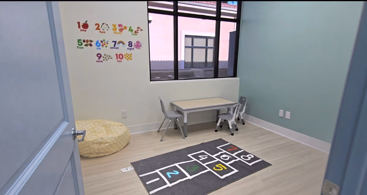 Pastel-walled therapy room with a hopscotch rug, small table, chairs, and a bean bag.