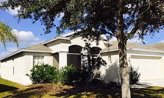 White single-story assisted living building with arched entrance, trees, and greenery.