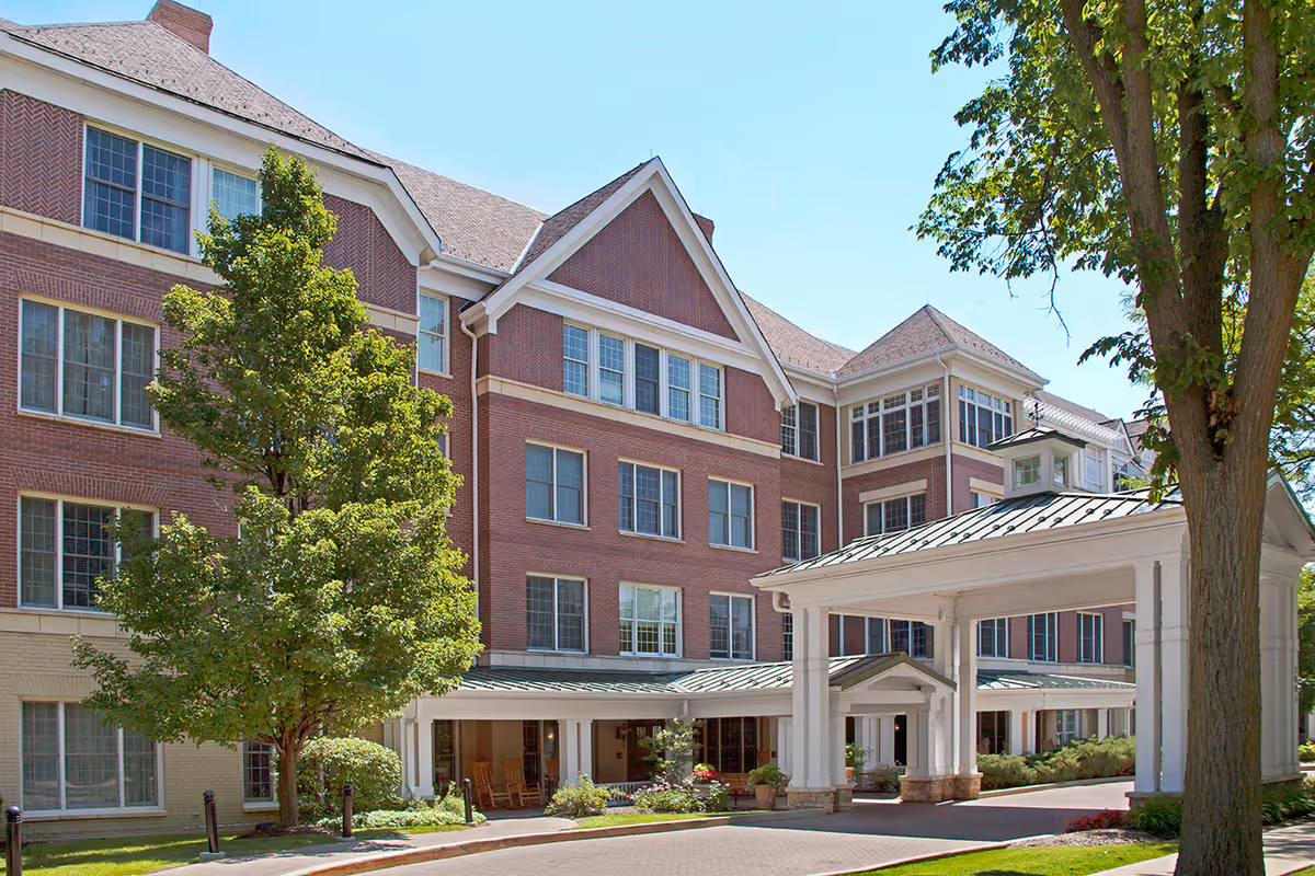 Brick senior living facility with a covered entrance, landscaping, and tall trees.