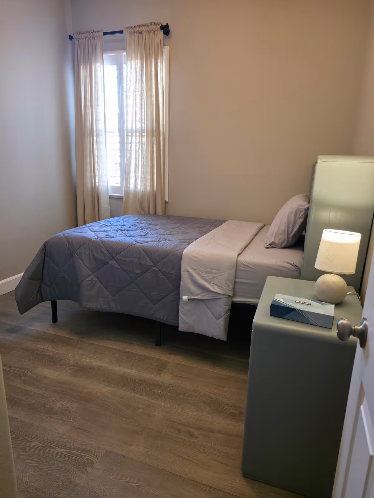 A simple bedroom in a senior living facility with a bed, nightstand, lamp, and window.