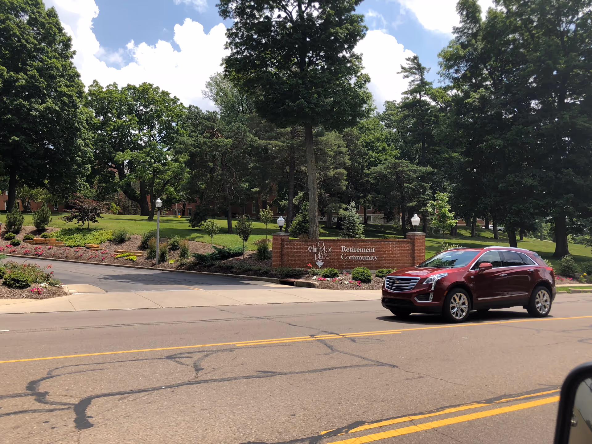 Brick entrance sign reads '10 Wilmington Place Retirement Community' with landscaped grounds and a passing car.