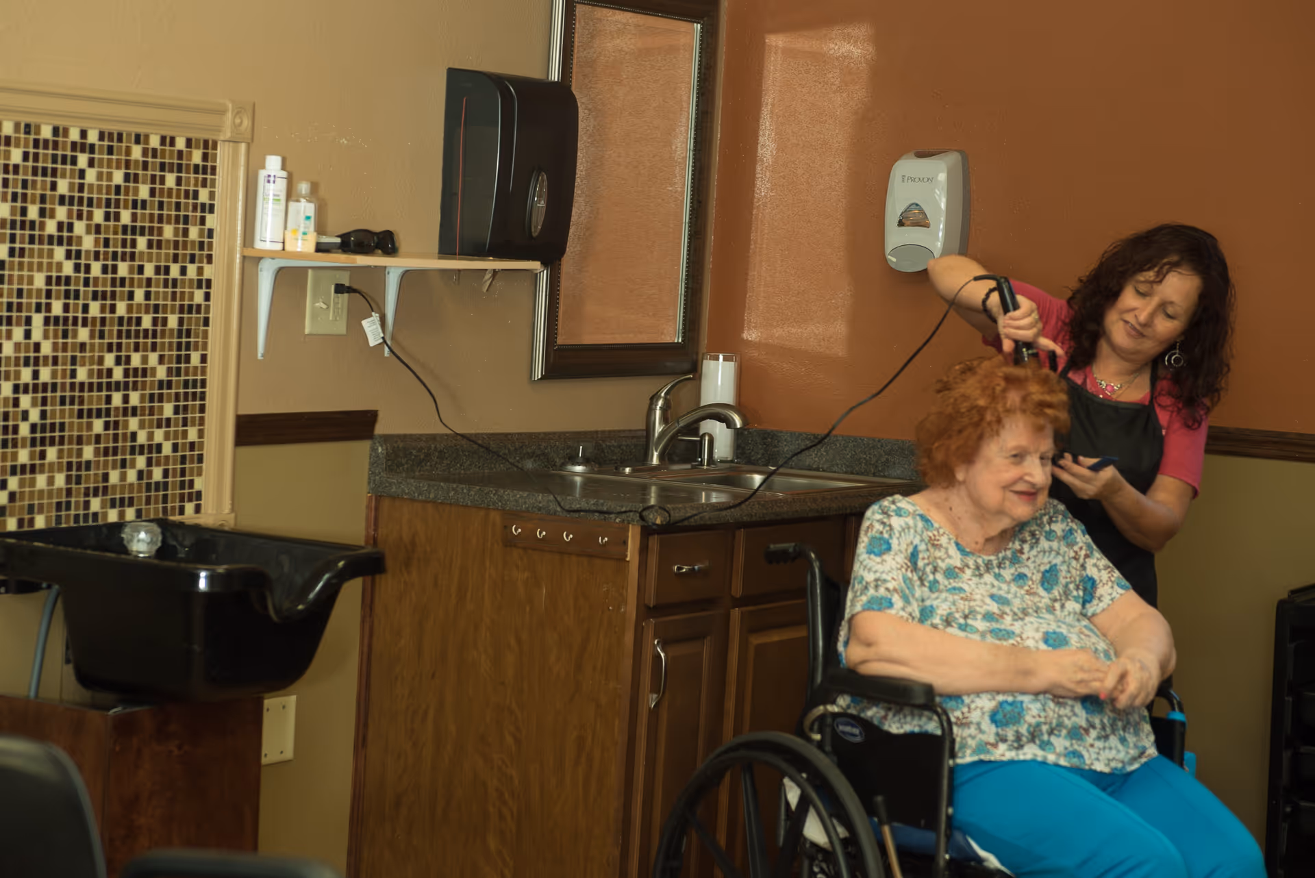 A woman in a wheelchair is getting her hair styled by a hairdresser in a salon area. The room has a sink for washing hair, a countertop with a mirror, and various hair care products on a shelf. The walls are painted in warm tones with a mosaic tile accent near the sink.