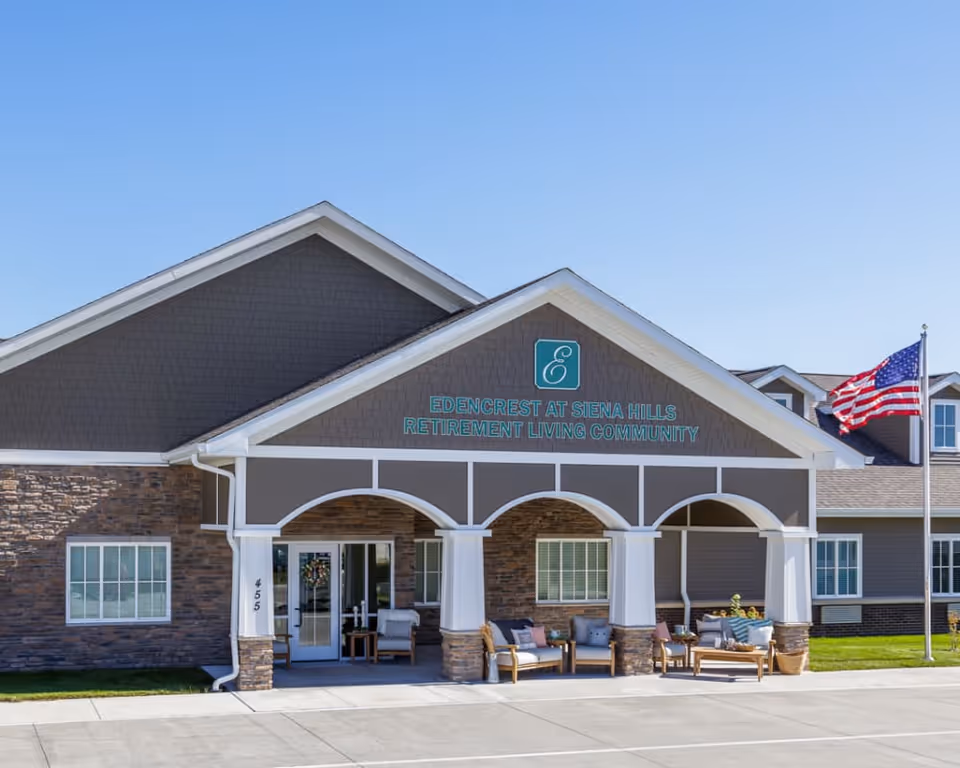 Front exterior view of Edencrest at Siena Hills retirement living community building with a covered entrance, outdoor seating area, and an American flag on a flagpole against a clear blue sky.