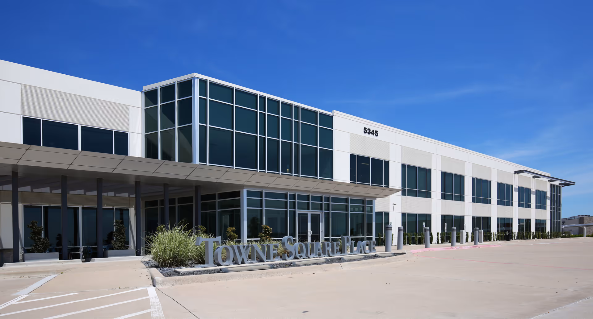 Modern glass-front building with a covered entrance and wide parking lot under blue sky.