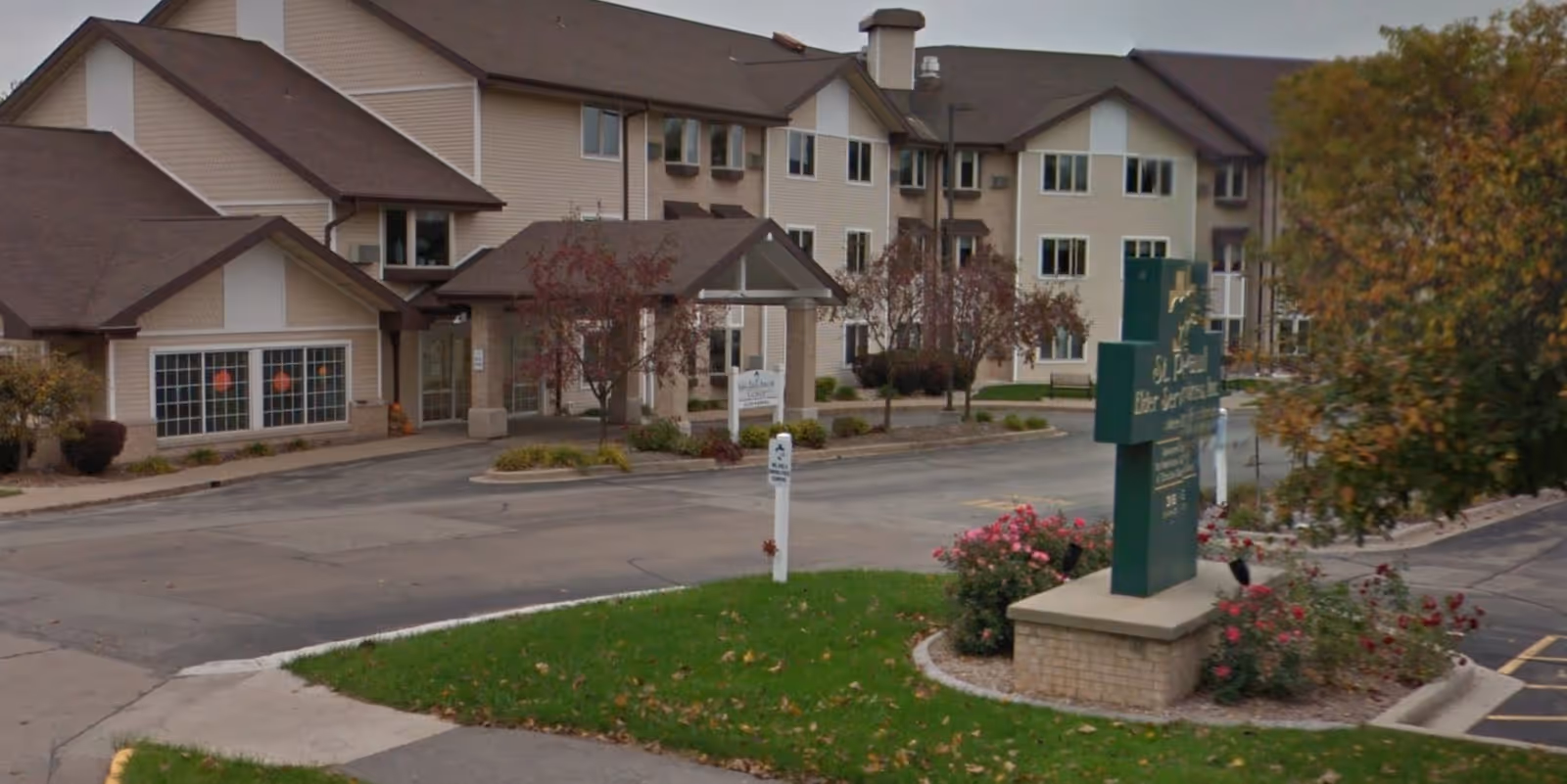 Exterior view of St Paul Villa, a multi-story senior living facility with beige siding and brown roofs. The entrance has a covered drop-off area with trees and landscaping around the driveway and parking lot.