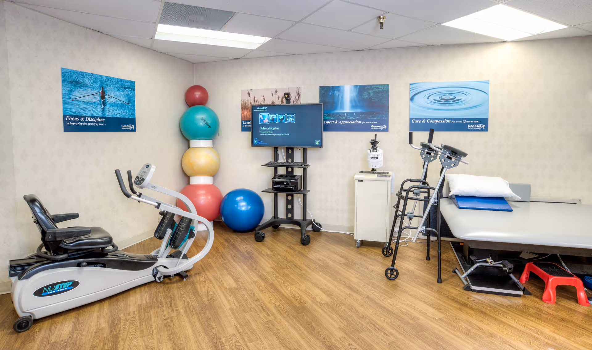 A rehabilitation therapy room with exercise equipment including a recumbent bike, colorful exercise balls stacked in a corner, a walker, a therapy table with a pillow and cushion, a TV mounted on a stand, and motivational posters on the walls.