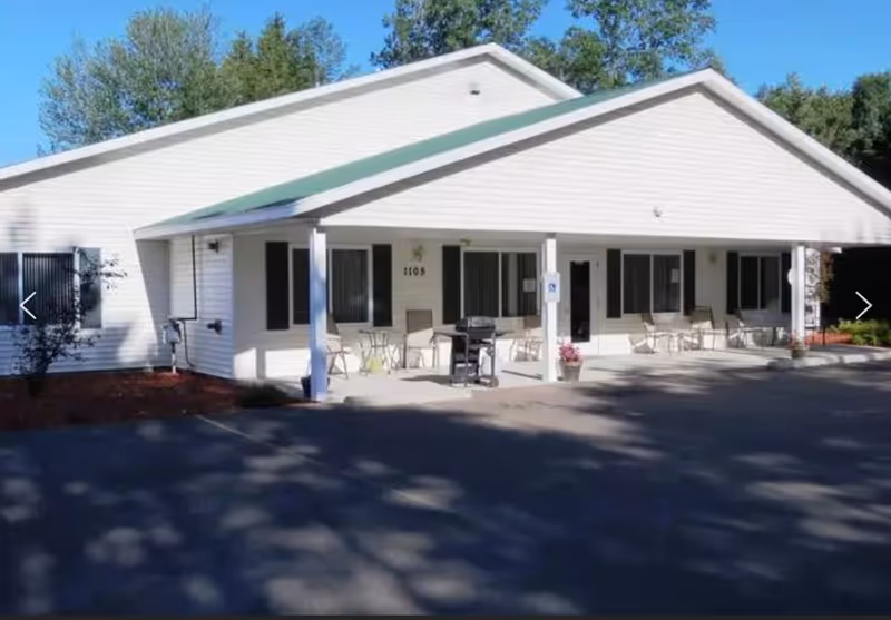 Exterior view of a single-story white building with a green roof and a covered porch area. The porch has several chairs, a table, a grill, and potted plants. Trees are visible in the background under a clear blue sky.