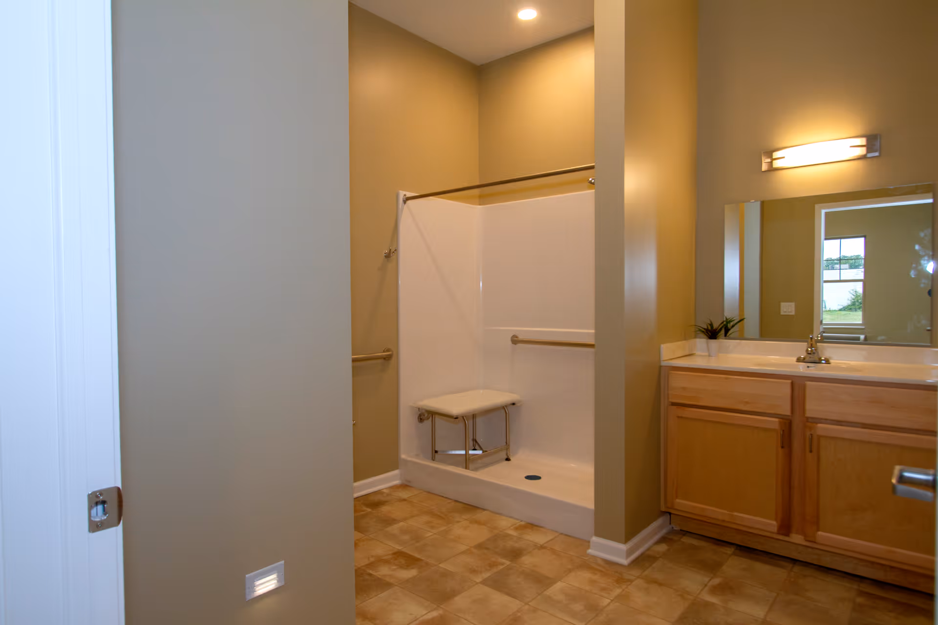 A bathroom with a walk-in shower featuring a built-in bench and grab bars. The room has beige walls, a tiled floor, a wooden vanity with a sink, and a large mirror above the vanity. A light fixture is mounted above the mirror.