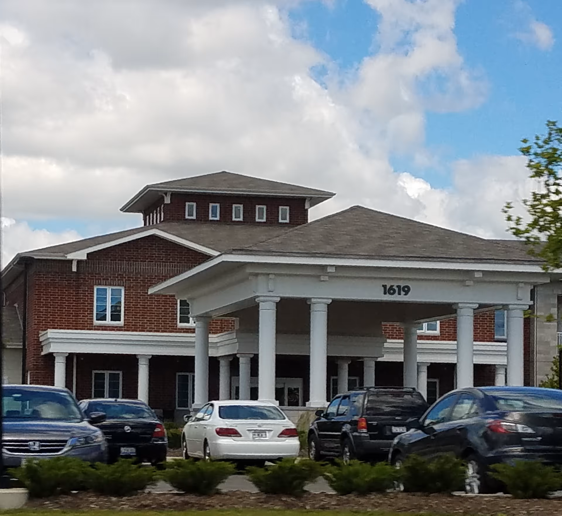 Exterior view of a brick senior living building with white portico and parked cars.