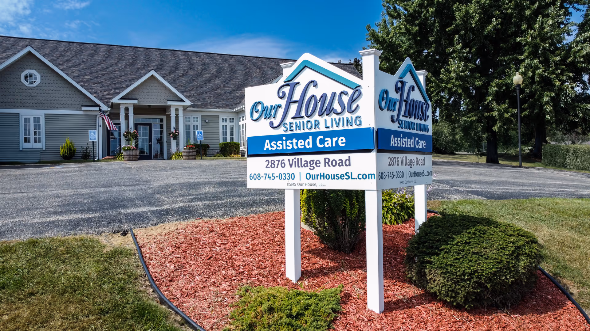 Front exterior of an assisted living building with a landscaped sign reading 'Our House Senior Living - Assisted Care' by the driveway.