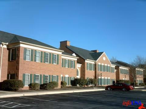 Two-story brick senior living building with green shutters, landscaped front, and parked cars under blue skies.