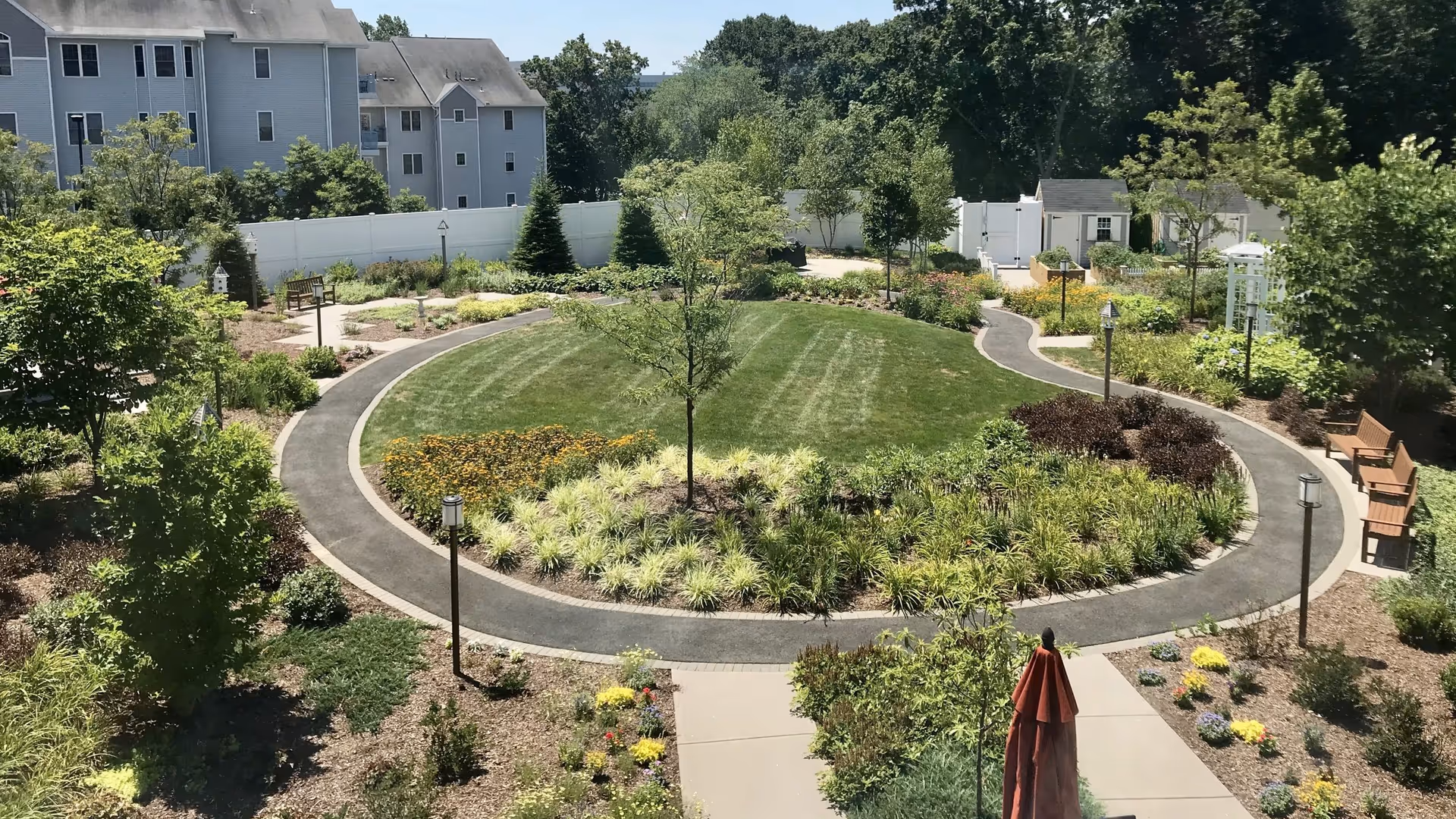 A well-maintained outdoor garden area with a circular walking path surrounding a grassy center with various plants and small trees. There are benches along the path, lamp posts, and a white fence enclosing the garden. In the background, there is a multi-story residential building and dense trees.
