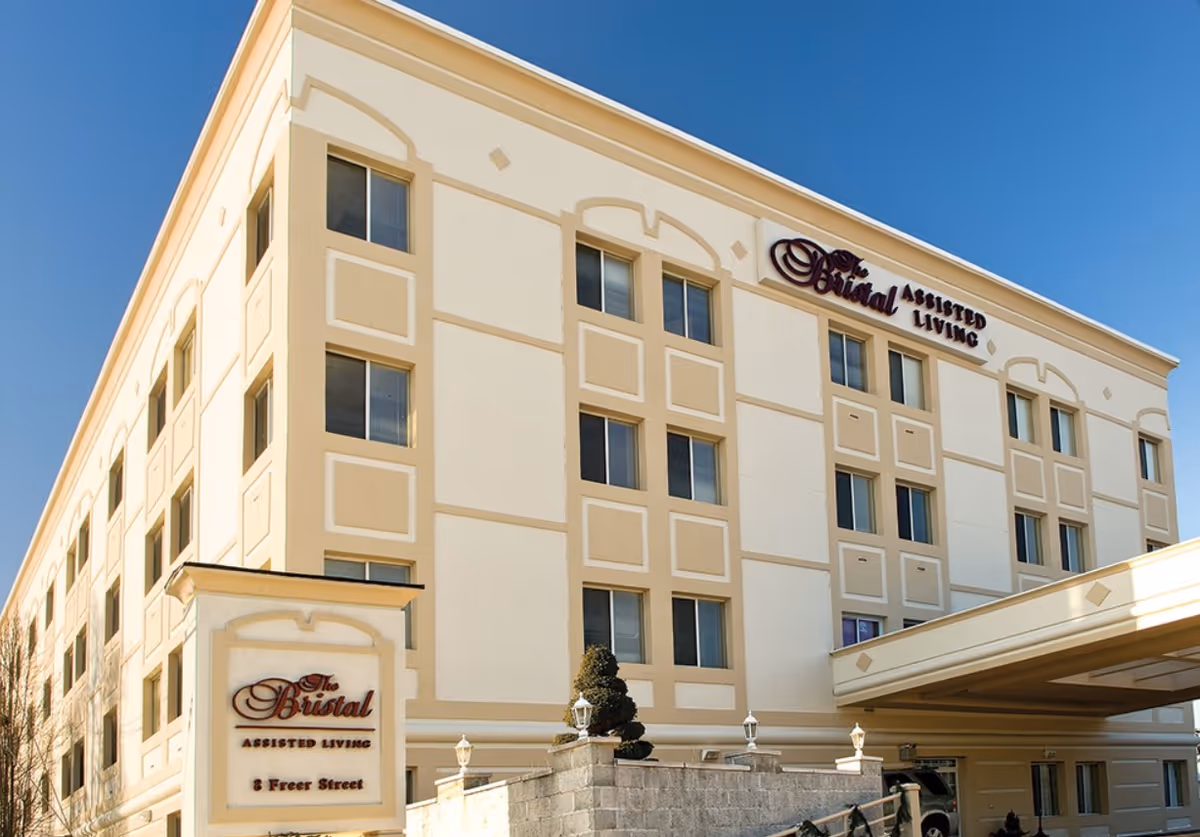 Front exterior of The Bristal Assisted Living building showing a beige-and-white multi-story facade, entrance canopy, and signage.