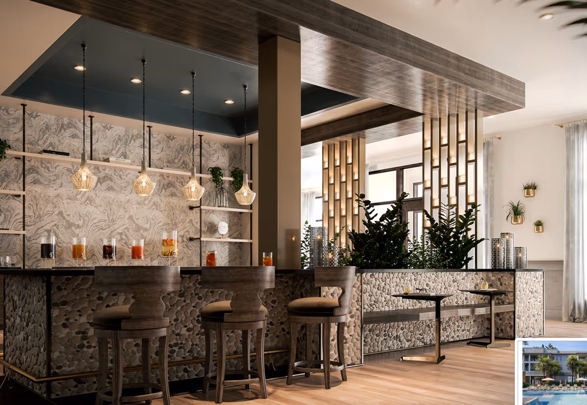 A stylish indoor bar area with a stone-textured counter and three wooden bar stools. Above the counter, there are four hanging pendant lights with glass shades. Behind the bar, shelves with decorative items and plants are mounted on a marbled wall. To the right, there are green plants and two small tables with chairs near large windows with light curtains, allowing natural light to fill the space.