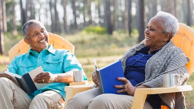 Two seniors smile and read books in outdoor chairs at a sunny facility courtyard.