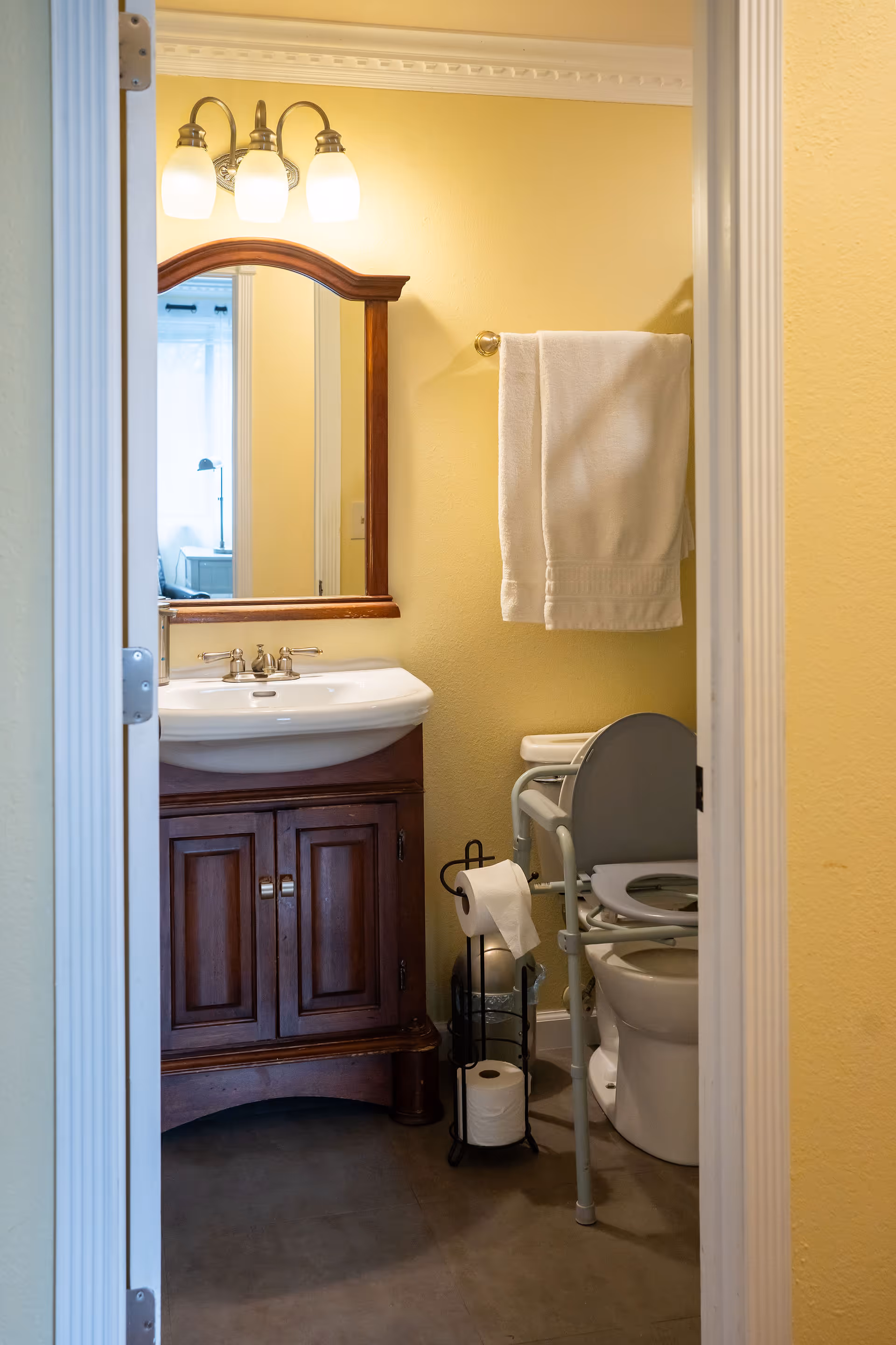 View into a small bathroom with yellow walls, featuring a wooden vanity with a white sink and a mirror above it. A three-light fixture is mounted above the mirror. To the right, there is a toilet with a raised seat and support rails, a towel hanging on a wall-mounted towel rack, and a toilet paper holder with extra rolls.
