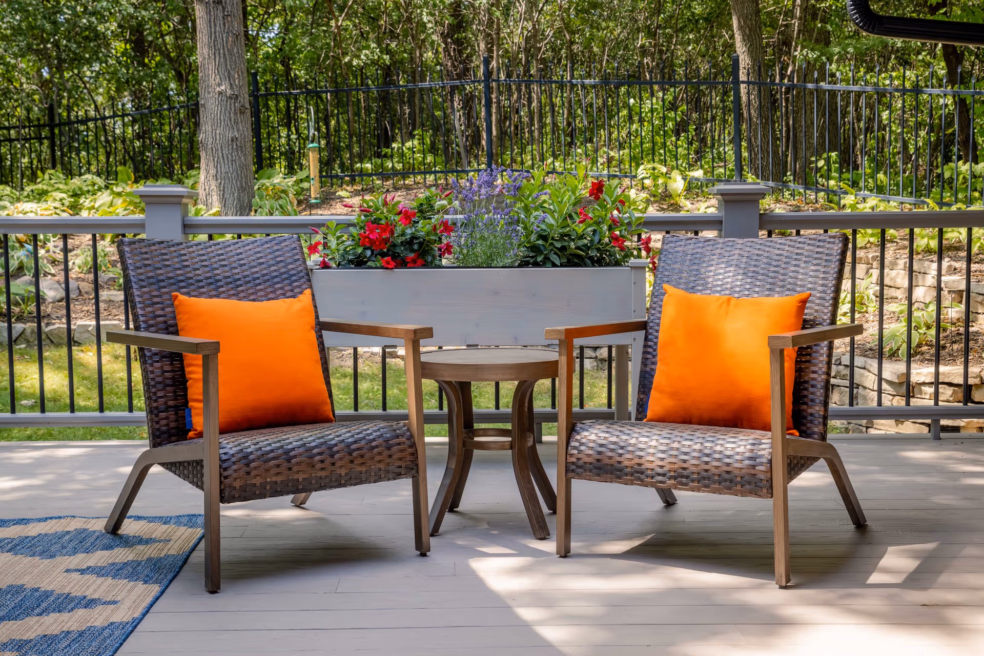 Outdoor patio area with two wicker chairs featuring bright orange cushions, a small round wooden table between them, and a planter box with red and purple flowers behind the table. The patio is surrounded by a black metal fence and lush green trees and plants.
