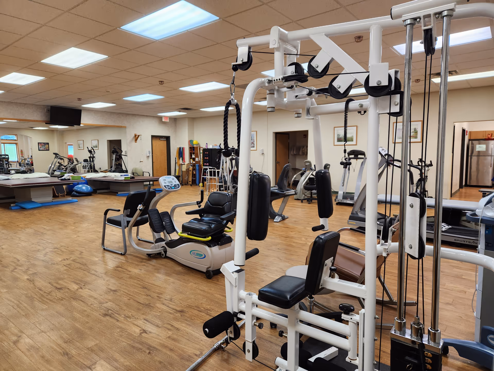 Well-lit gym at Cortland Acres Nursing Home with weight machines, treadmills, and wooden flooring.