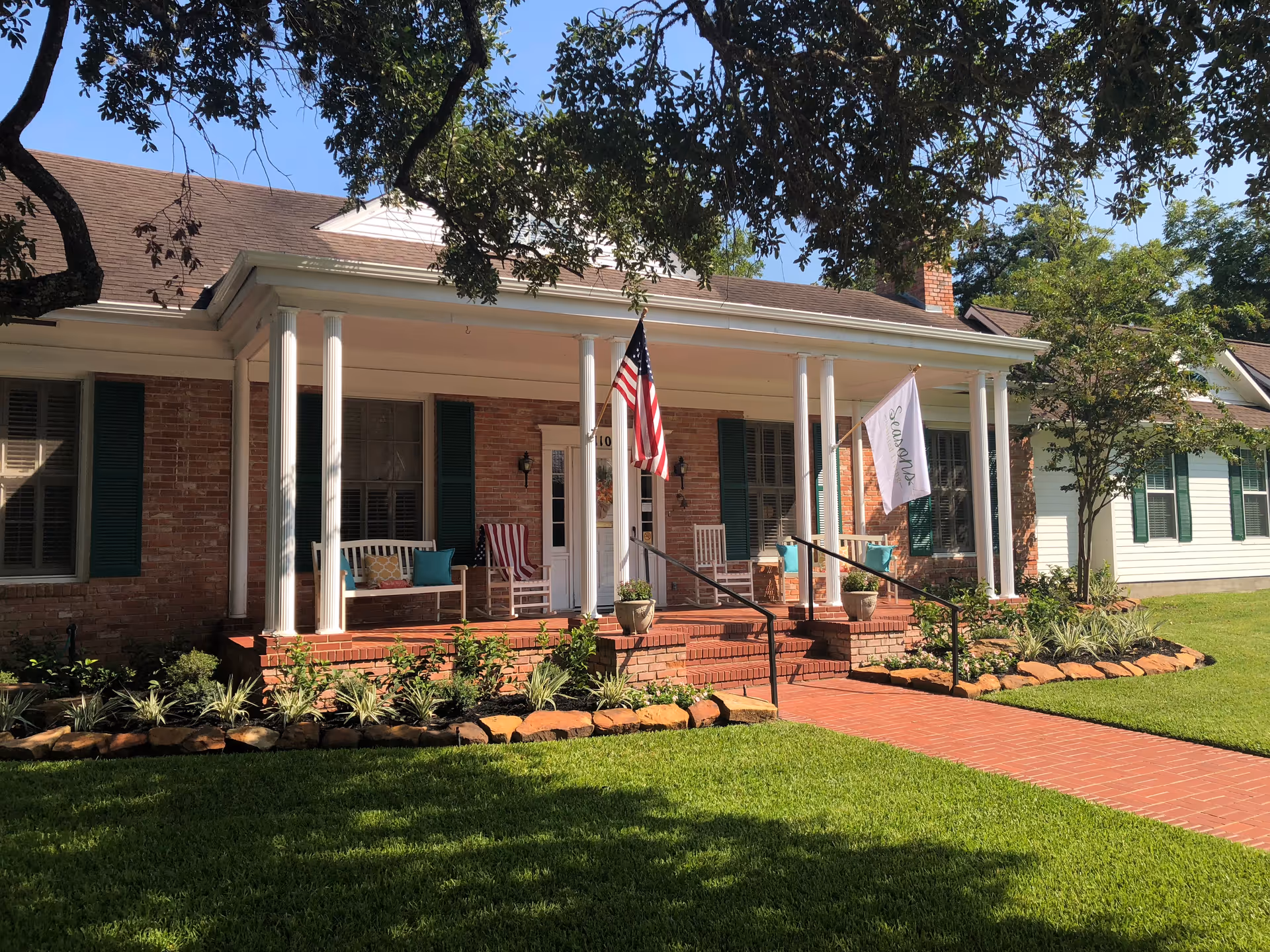 Front porch of a brick assisted living facility with columns, rocking chairs, flags, and lawn.