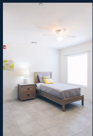 Bright bedroom in a senior living facility featuring a single bed, nightstand, lamp, and window.