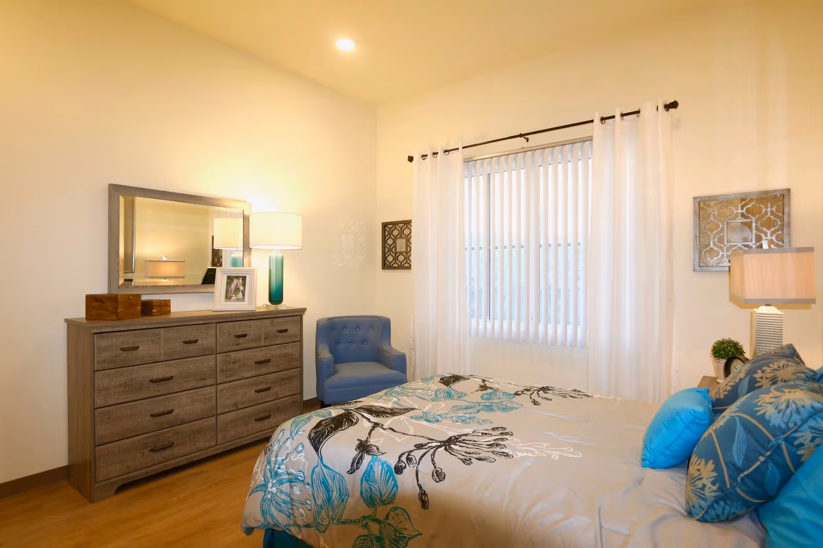 A cozy bedroom with a bed covered in a floral-patterned comforter and blue accent pillows. There is a wooden dresser with a mirror above it, a table lamp, and a framed photo. A blue upholstered chair sits in the corner near a window with vertical blinds and white curtains. The room has light-colored walls and wooden flooring.