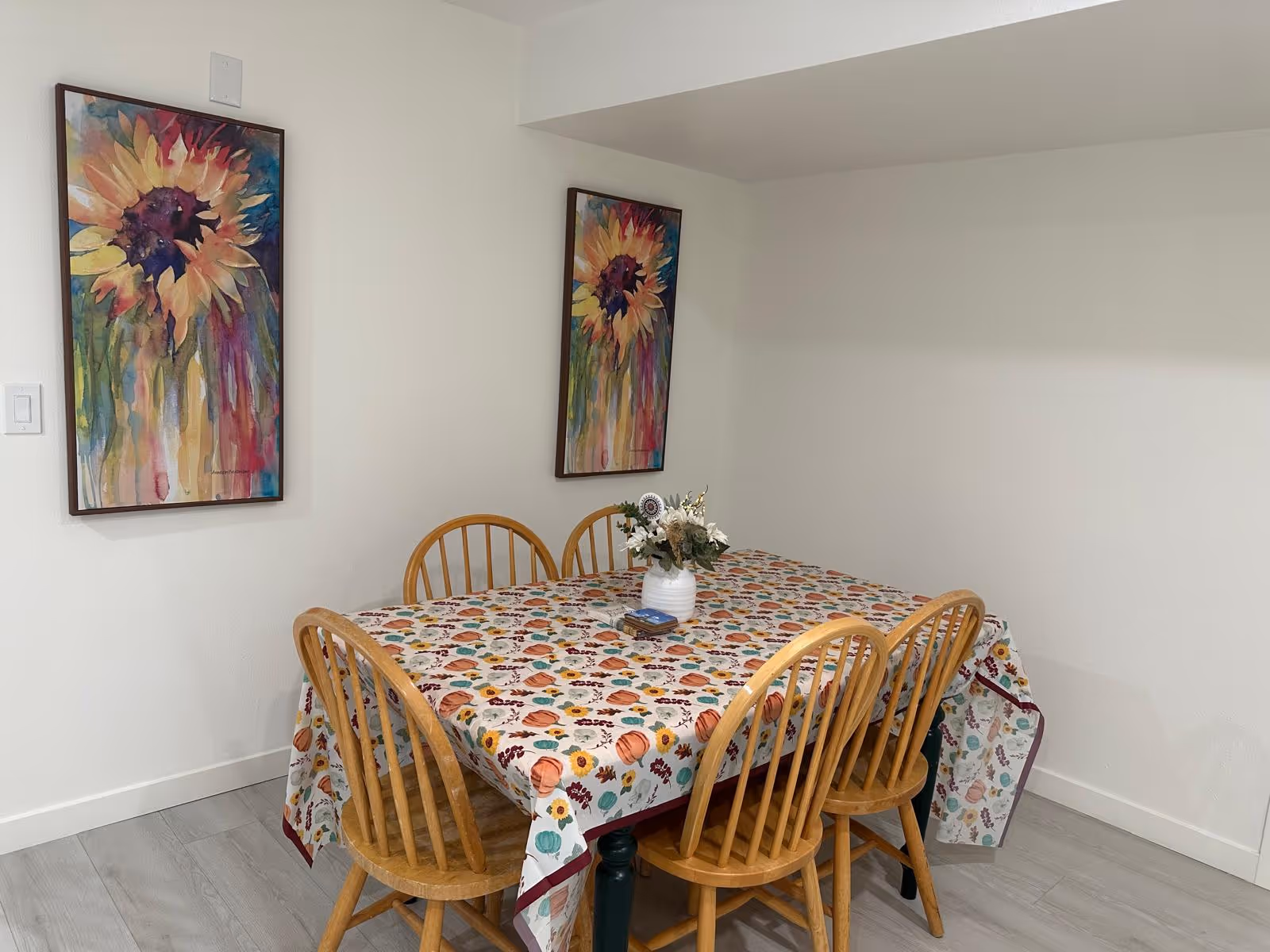 A bright dining room with a patterned tablecloth, six wooden chairs, and colorful wall art.