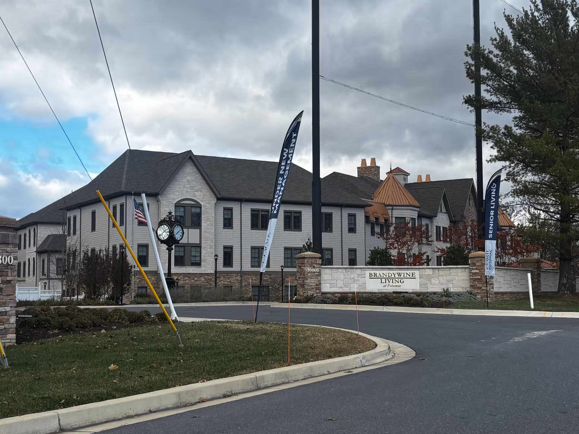 Exterior view of Brandywine Living At Potomac, a senior living facility with a large multi-story building, a clock on a post, American flag, and banners near the entrance. The sky is cloudy with patches of blue.