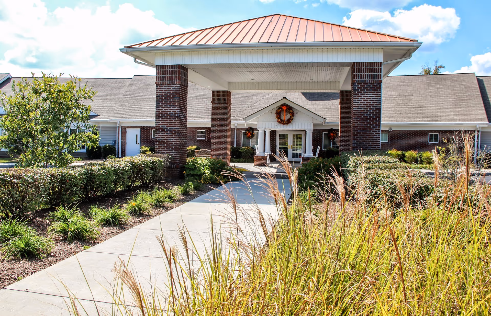 Front entrance of a brick building with a covered walkway supported by brick columns. The building has a red metal roof and a wreath decoration above the door. There are landscaped bushes and tall grasses along the concrete pathway leading to the entrance under a partly cloudy sky.