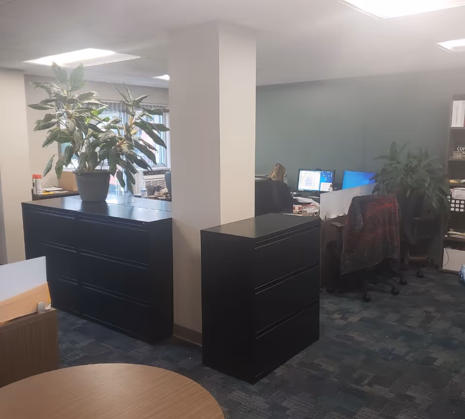 Office area with desks, filing cabinets, and a person at a computer near a pillar.