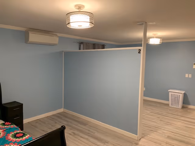 Blue-walled bedroom with partitioned cubicles, bed, dresser, and laundry basket.