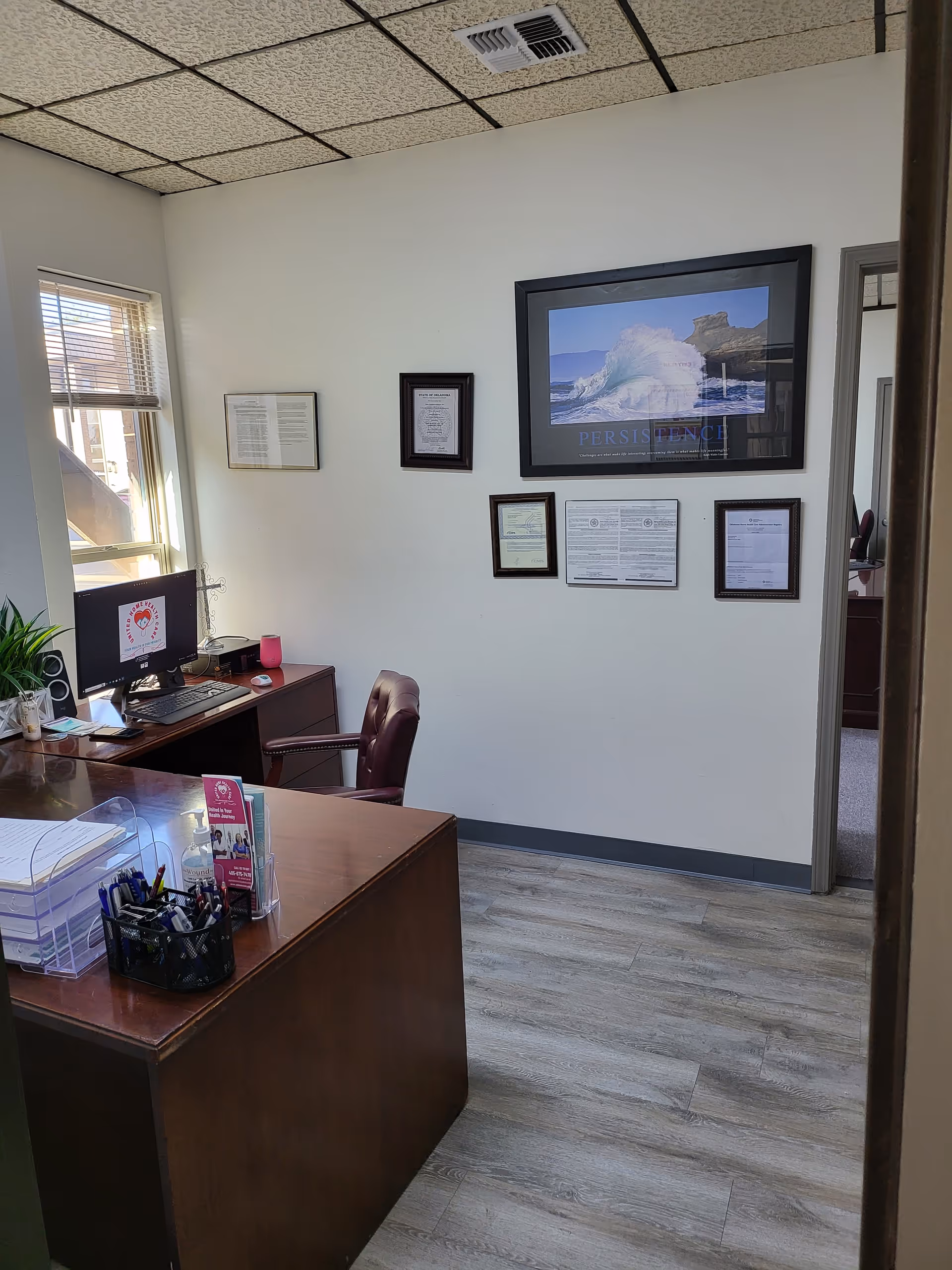 Office desk with computer, chairs, and framed certificates on a beige wall.
