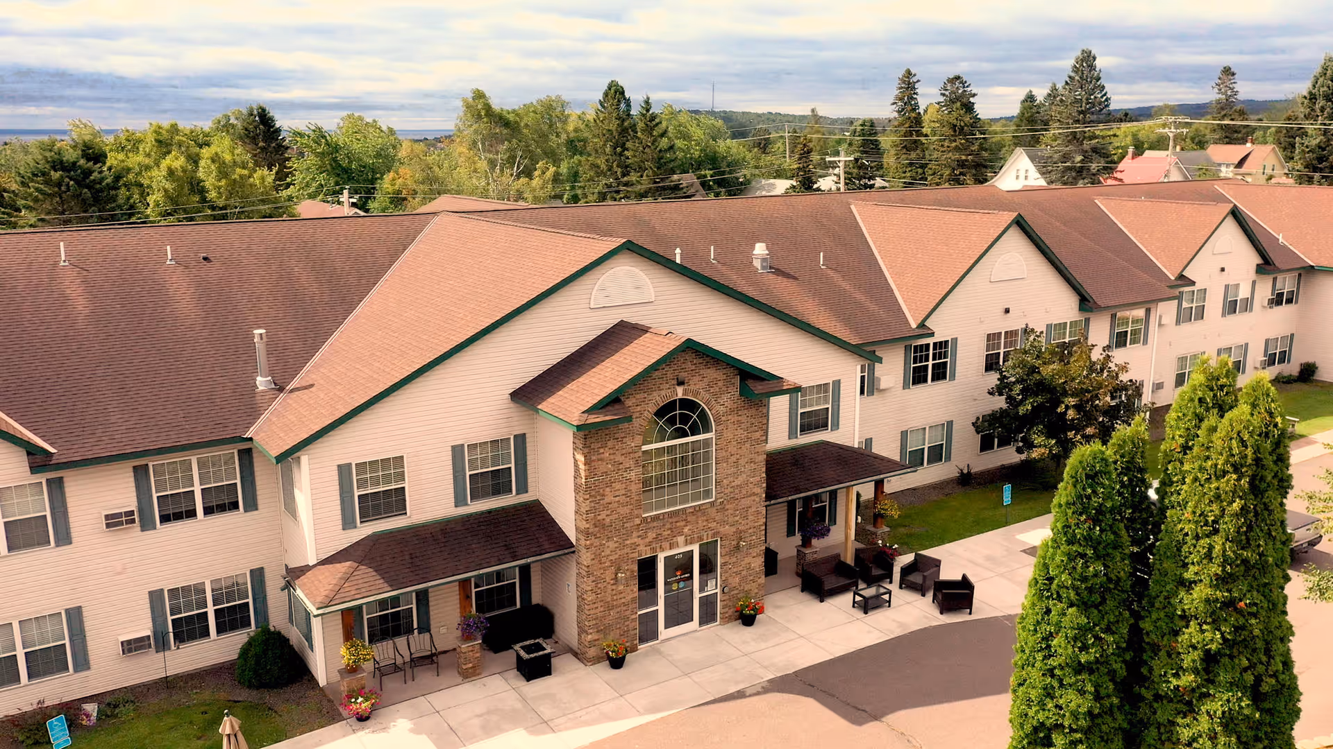 Exterior view of a two-story senior living facility building with a brown roof and beige siding. The entrance features a brick facade with large windows above the door. There are outdoor seating areas with chairs and tables near the entrance, surrounded by greenery and tall trees. The sky is partly cloudy.