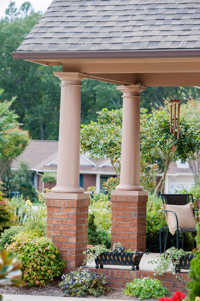 A covered outdoor patio area with two beige columns on brick bases, surrounded by lush green bushes and plants. There is a black metal chair with a beige cushion and a hanging wind chime. In the background, there are trees and a house with a sloped roof.