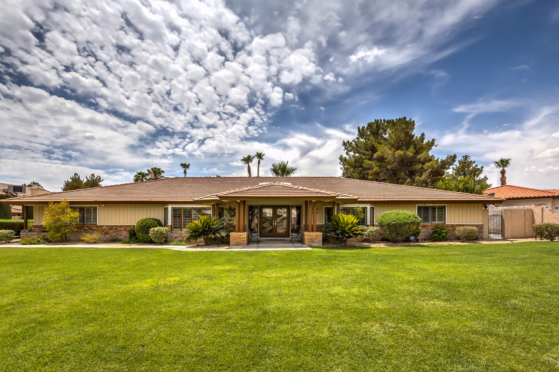 Front exterior view of a single-story building with a tiled roof, beige siding, and brick accents. The building is surrounded by a well-maintained green lawn, various shrubs, and palm trees under a partly cloudy sky.