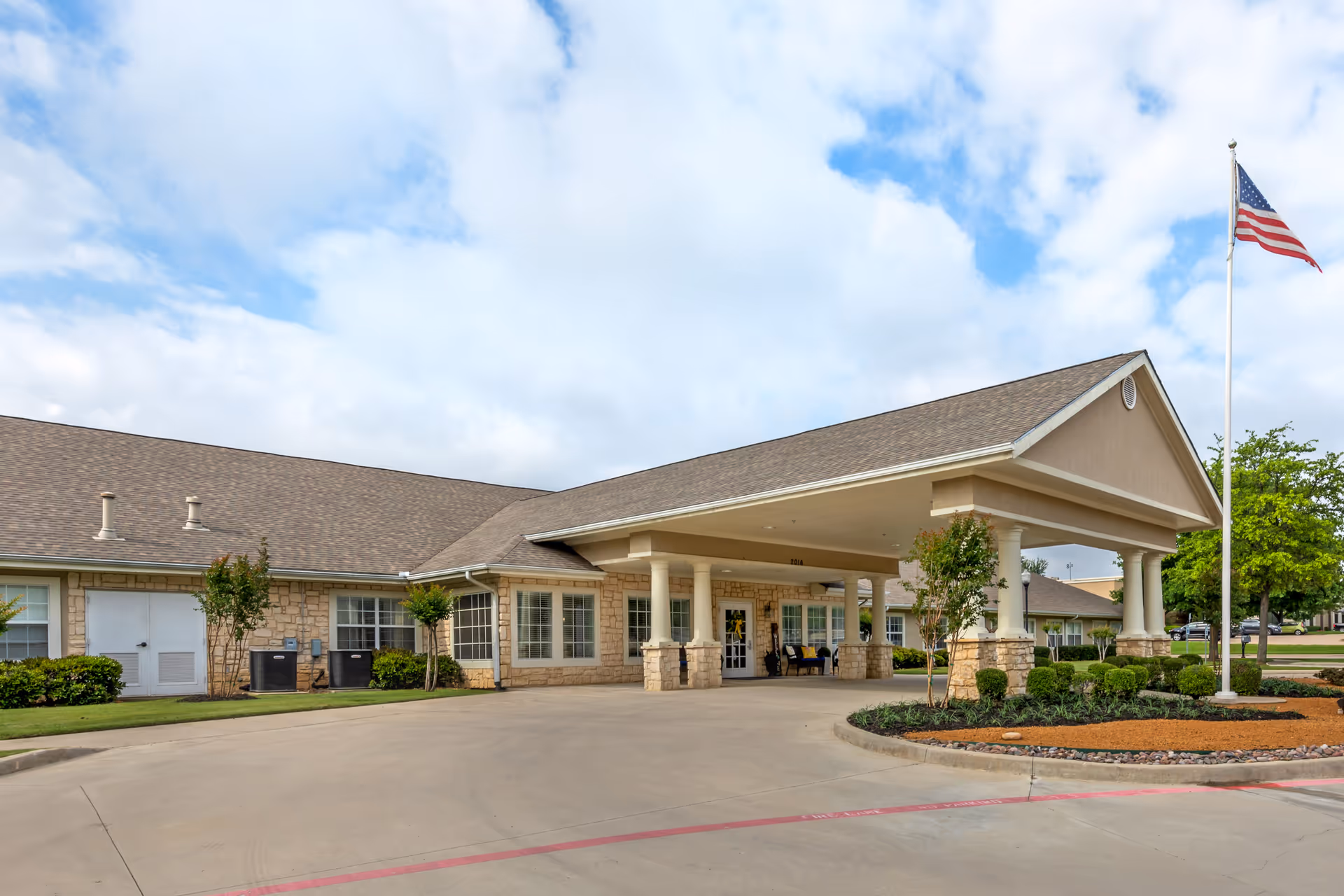 Single-story senior living facility with a covered entrance, drive-up circle, and American flag.