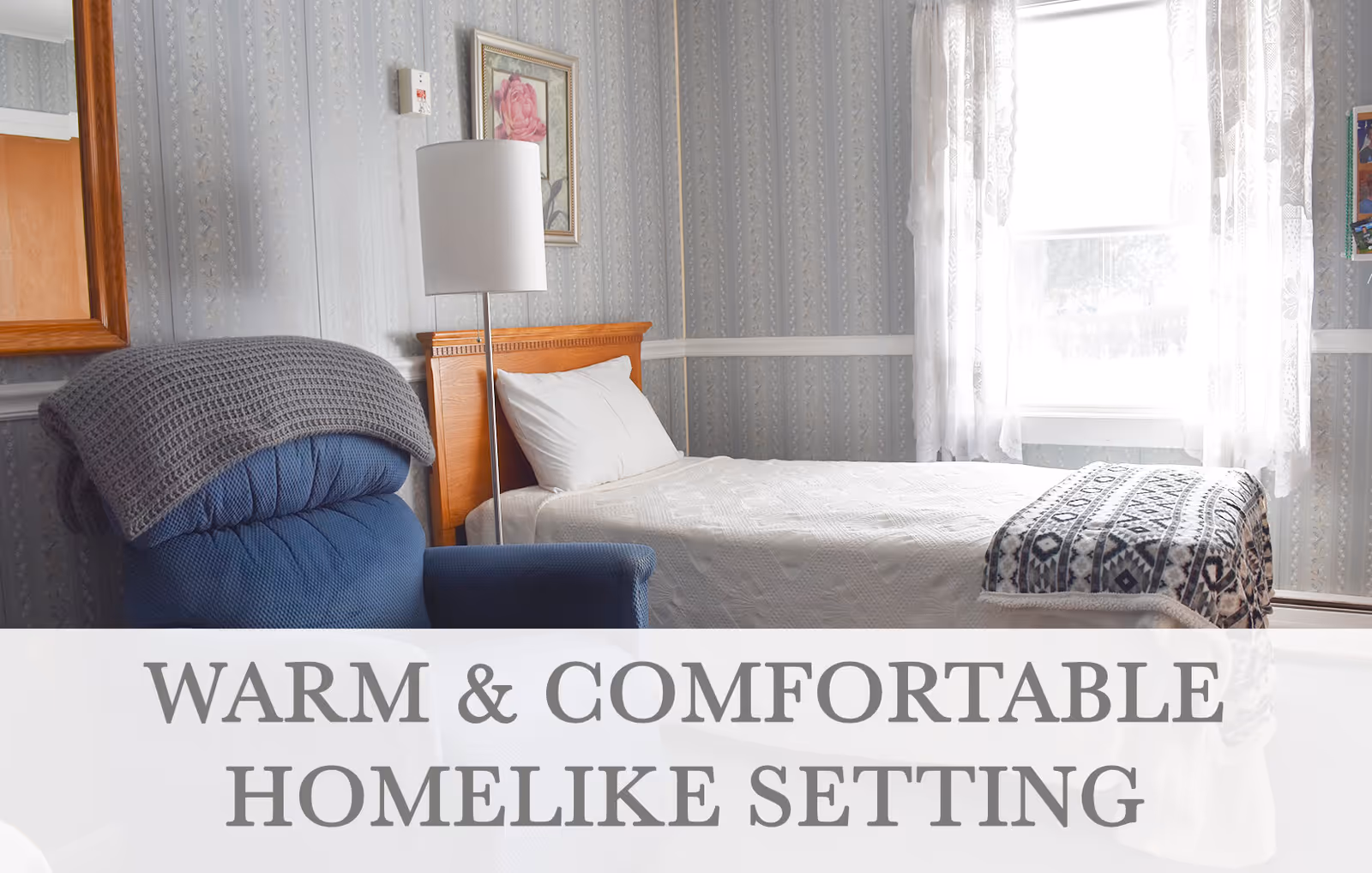 A cozy bedroom with a single bed covered in a white quilt and a patterned blanket at the foot. Next to the bed is a blue armchair with a gray knitted blanket draped over it. A floor lamp stands between the chair and the bed. The room has light gray patterned wallpaper, a window with sheer white curtains letting in natural light, and a framed picture of a pink rose on the wall.
