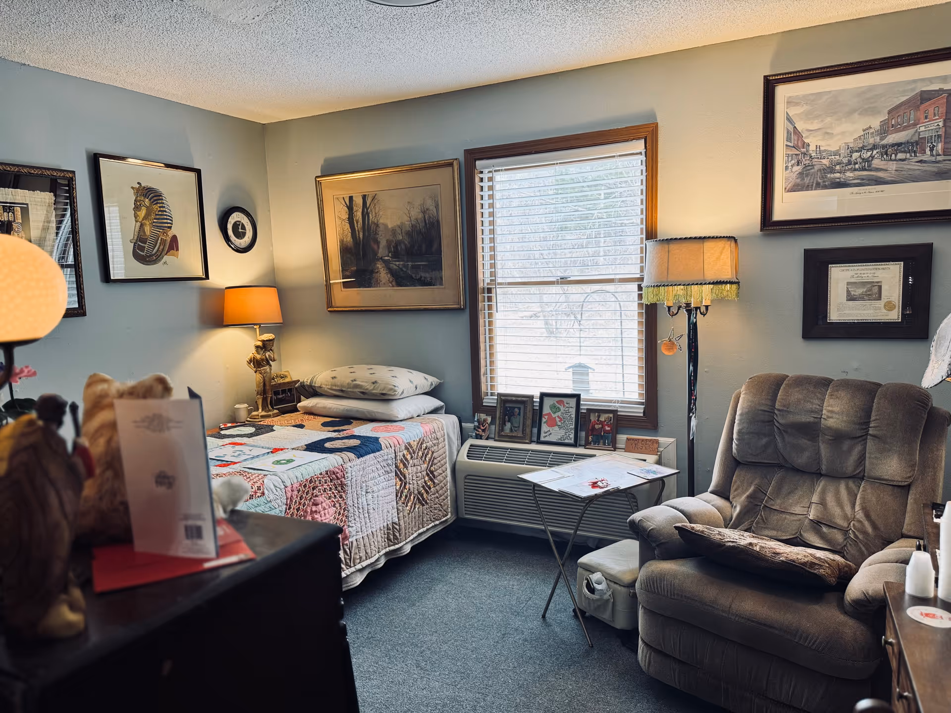 Cozy senior living bedroom with quilted bed, recliner, lamps, and framed artwork.