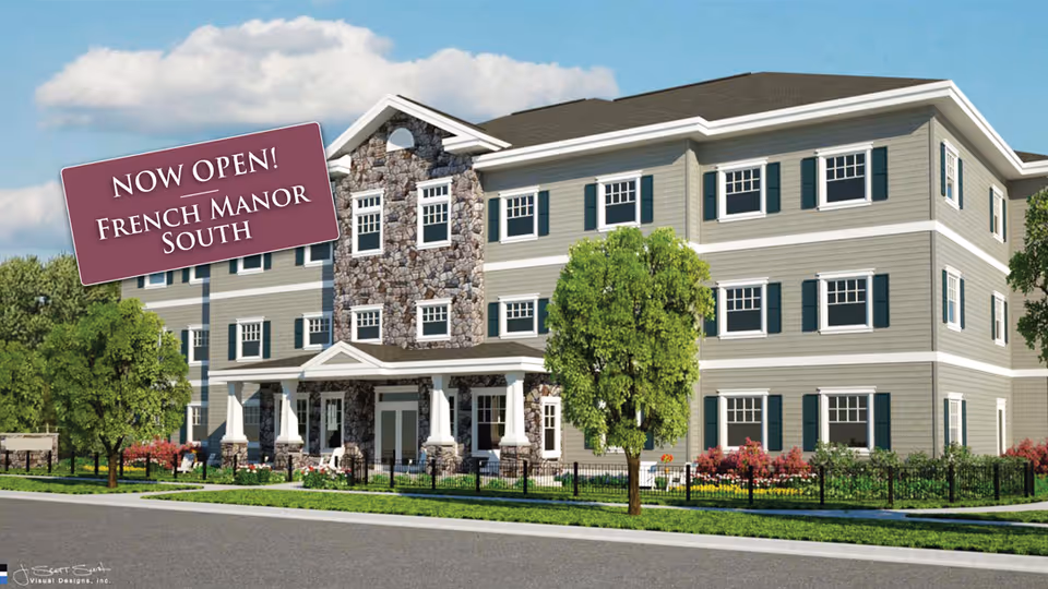 Exterior view of a three-story senior living facility building with stone and siding facade, surrounded by trees, shrubs, and a black metal fence under a blue sky with clouds.
