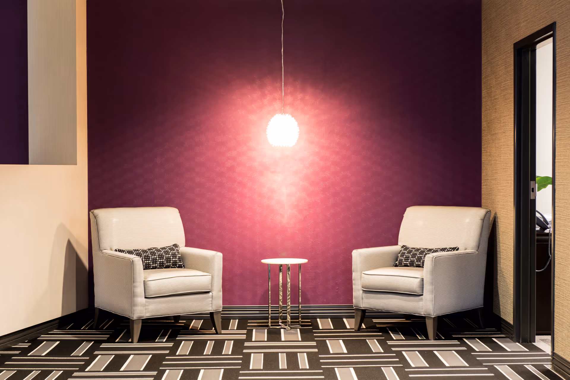 Two white armchairs with patterned cushions facing each other with a small round table between them, set against a purple wall with a hanging light fixture above the table. The floor has a geometric black and white pattern, and there is a doorway visible on the right side.