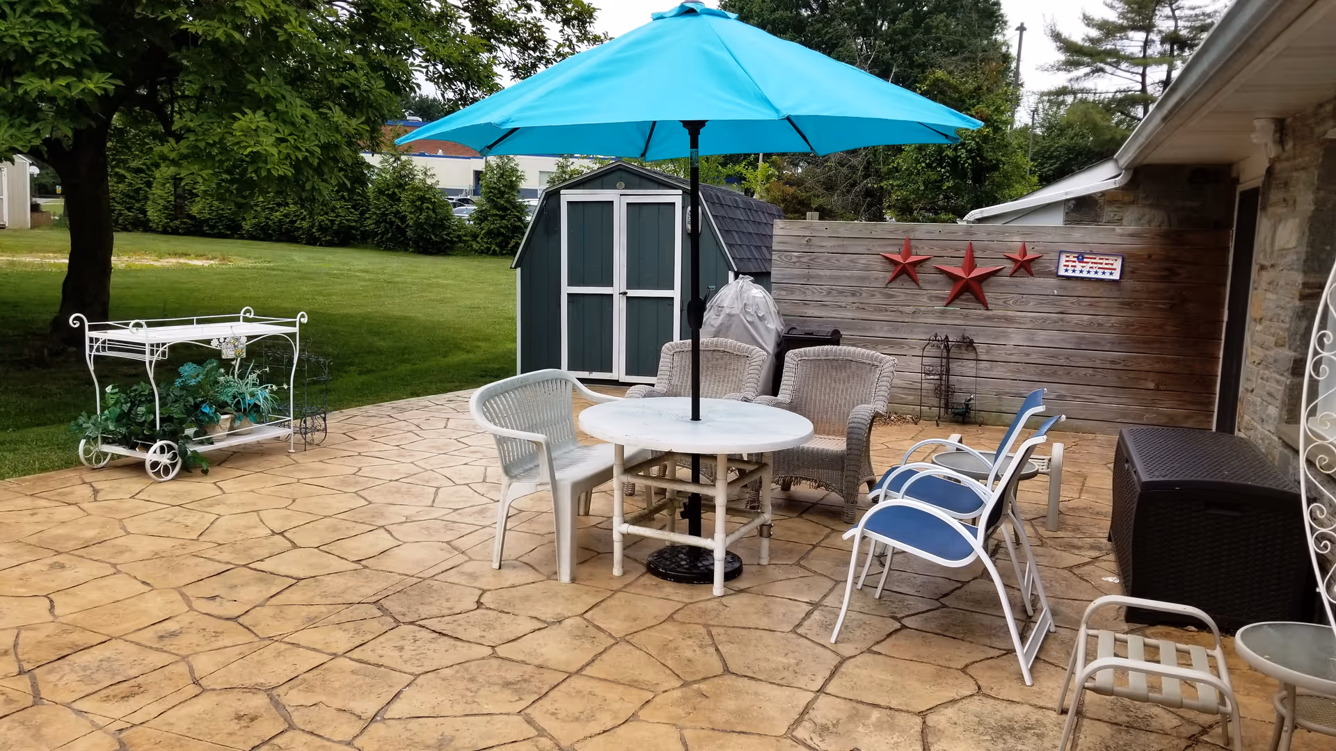Outdoor patio area with a round table and a blue umbrella surrounded by various chairs including white plastic and wicker chairs. There is a small white metal plant stand with green plants on the left side, a green shed in the background, and a wooden fence decorated with three red stars and a small American flag plaque. The patio is made of stamped concrete and is adjacent to a stone building wall.