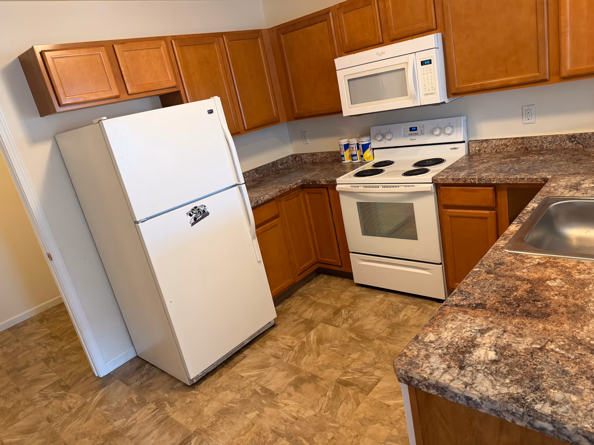 Bright U-shaped kitchen with white refrigerator, stove, microwave, granite countertops, and wood cabinets.