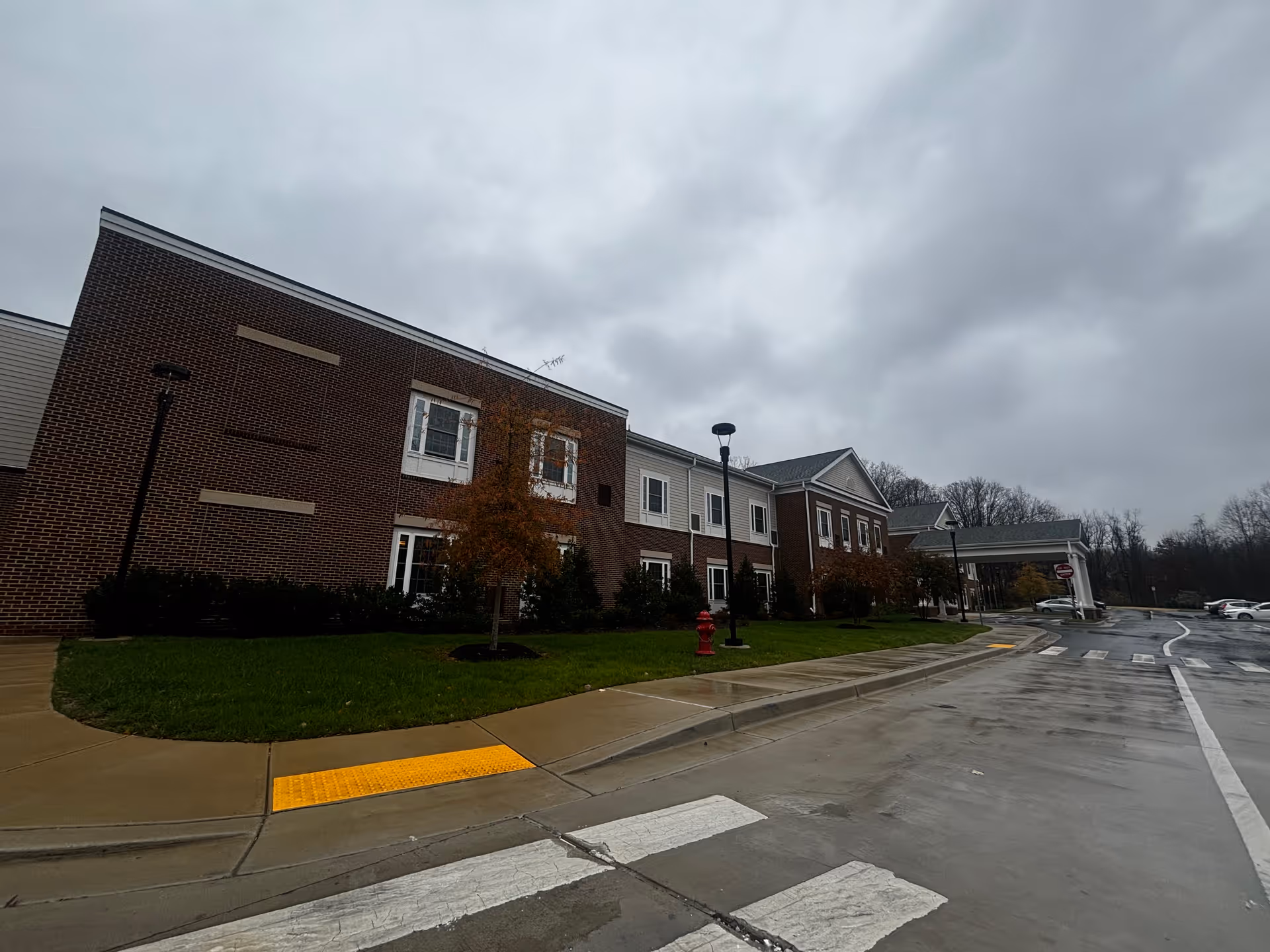 Exterior view of a two-story brick and siding building on a cloudy day with a wet driveway and sidewalk. There are small trees and bushes along the building, a red fire hydrant, street lamps, and a covered entrance with a 'Do Not Enter' sign. Several parked cars are visible in the background.