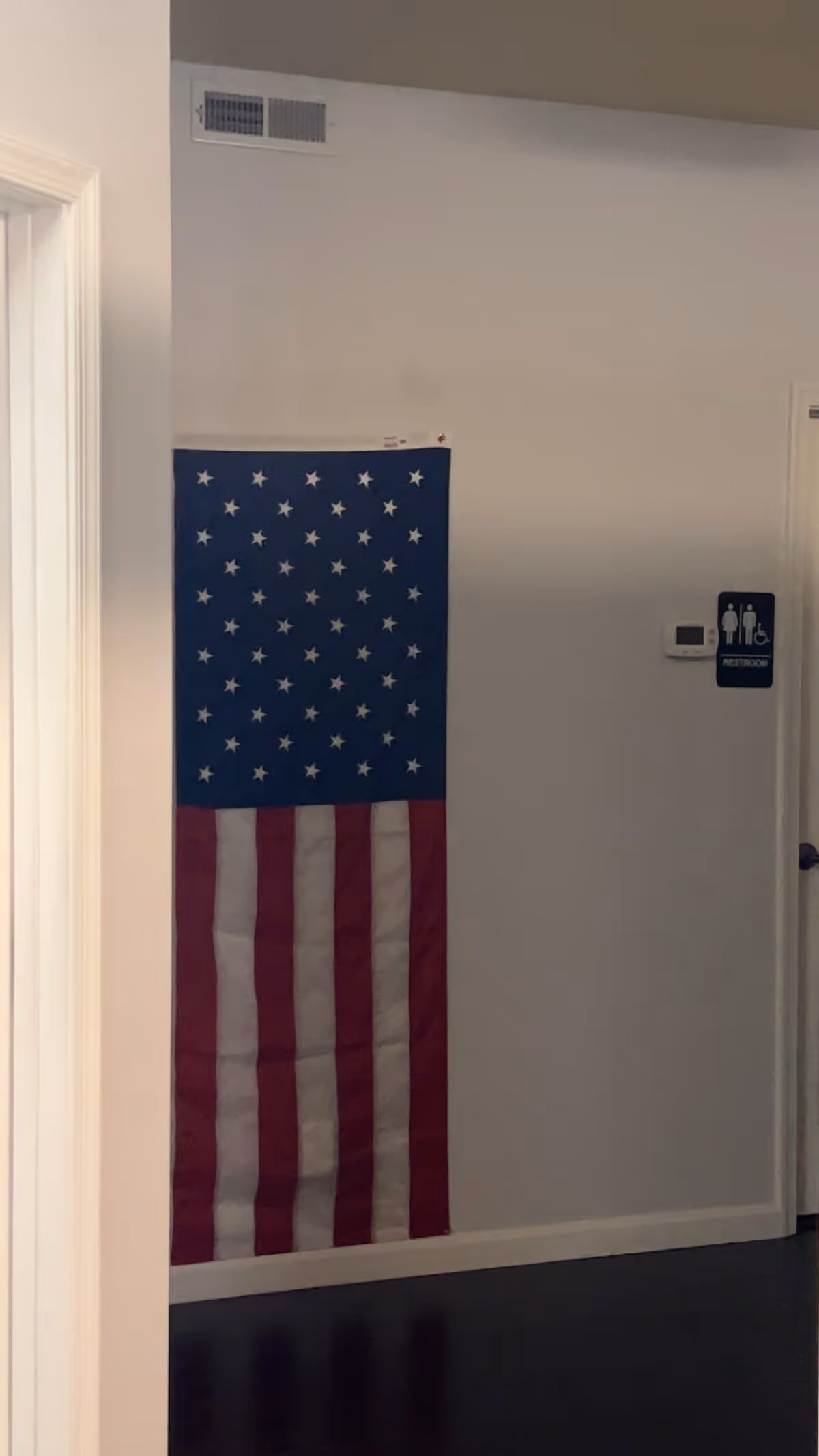 Tall American flag hangs on a white wall inside a facility hallway near a restroom sign.