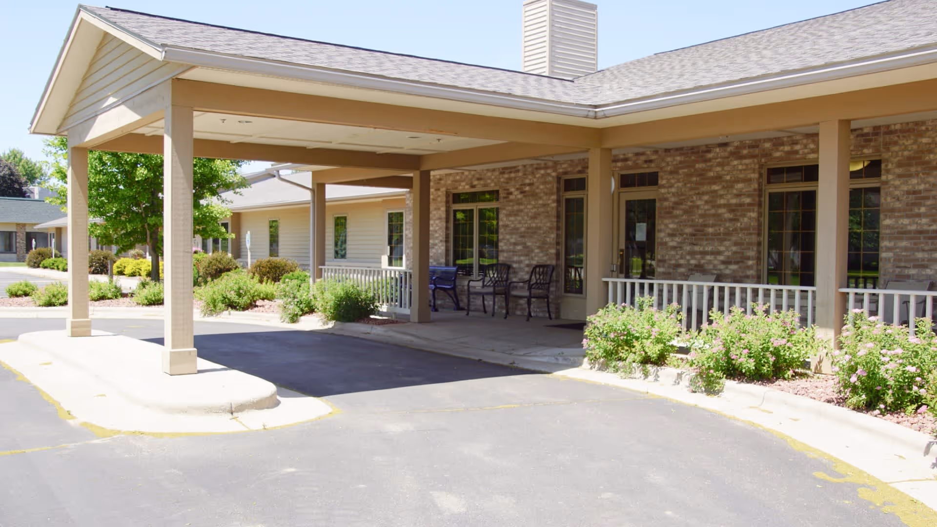 Covered entrance area of a senior living facility with a beige brick exterior, several chairs on the porch, and surrounding greenery including bushes and trees under a clear sky.
