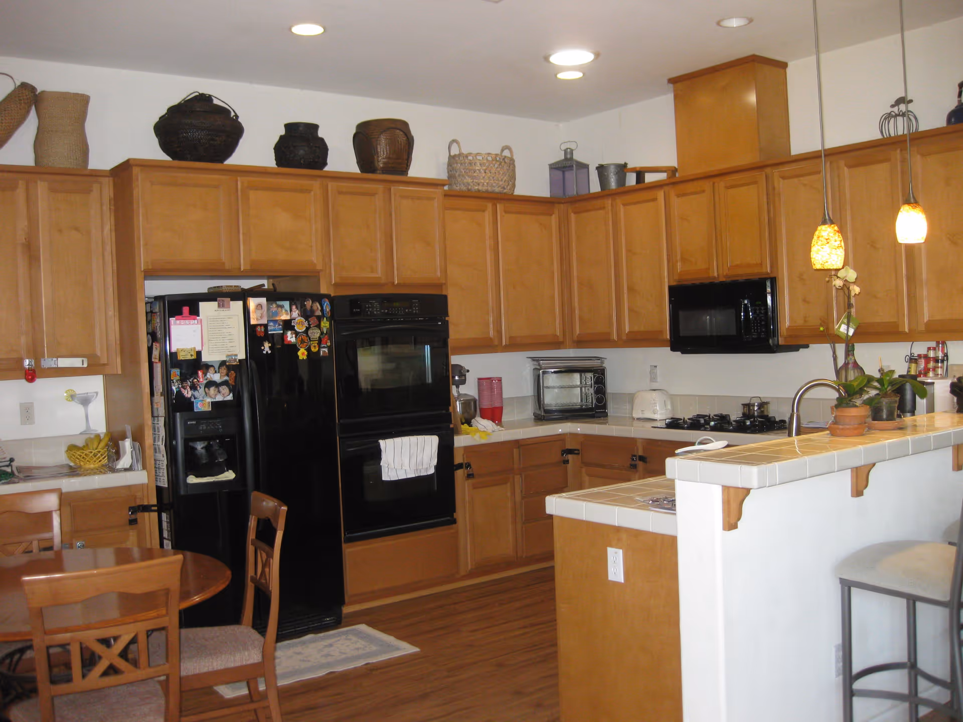 A kitchen with wooden cabinets, a black refrigerator covered with photos and magnets, a double oven, a microwave, a toaster, a toaster oven, and a gas stove. There is a small round dining table with chairs in the foreground and a counter with two pendant lights and bar stools on the right side. Various baskets and decorative items are placed on top of the cabinets.