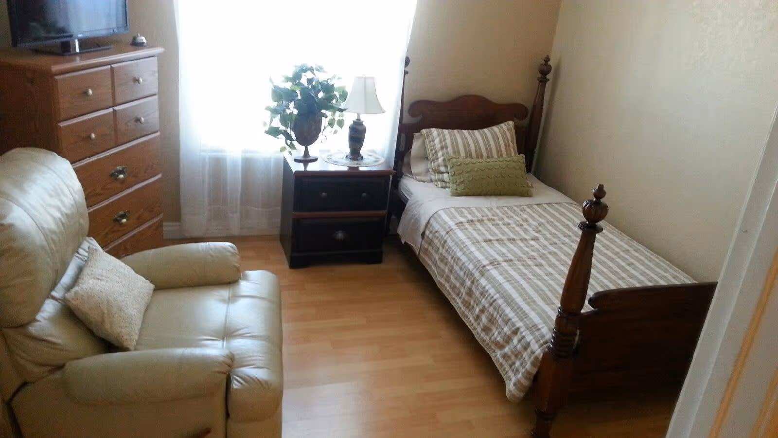 A small bedroom with a single wooden bed with striped bedding and pillows, a beige leather armchair with a cushion, a wooden dresser with a TV on top, and a nightstand with a lamp and a potted plant in front of a window with sheer white curtains.