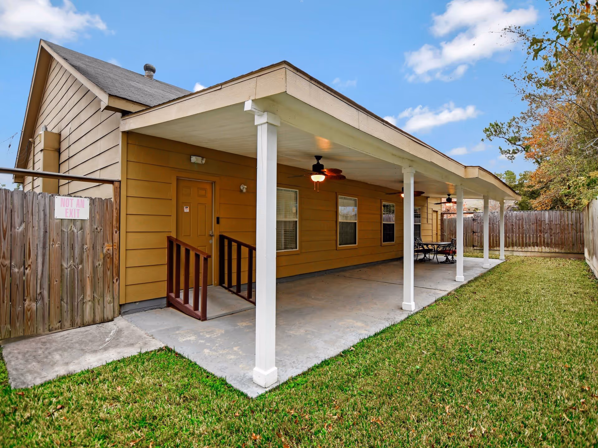 Outdoor covered patio area attached to a yellow building with white pillars supporting the roof. The patio has ceiling fans with lights and a table with chairs at the far end. The area is enclosed by a wooden fence with a sign that reads 'NOT AN EXIT'. There is a grassy lawn adjacent to the patio.