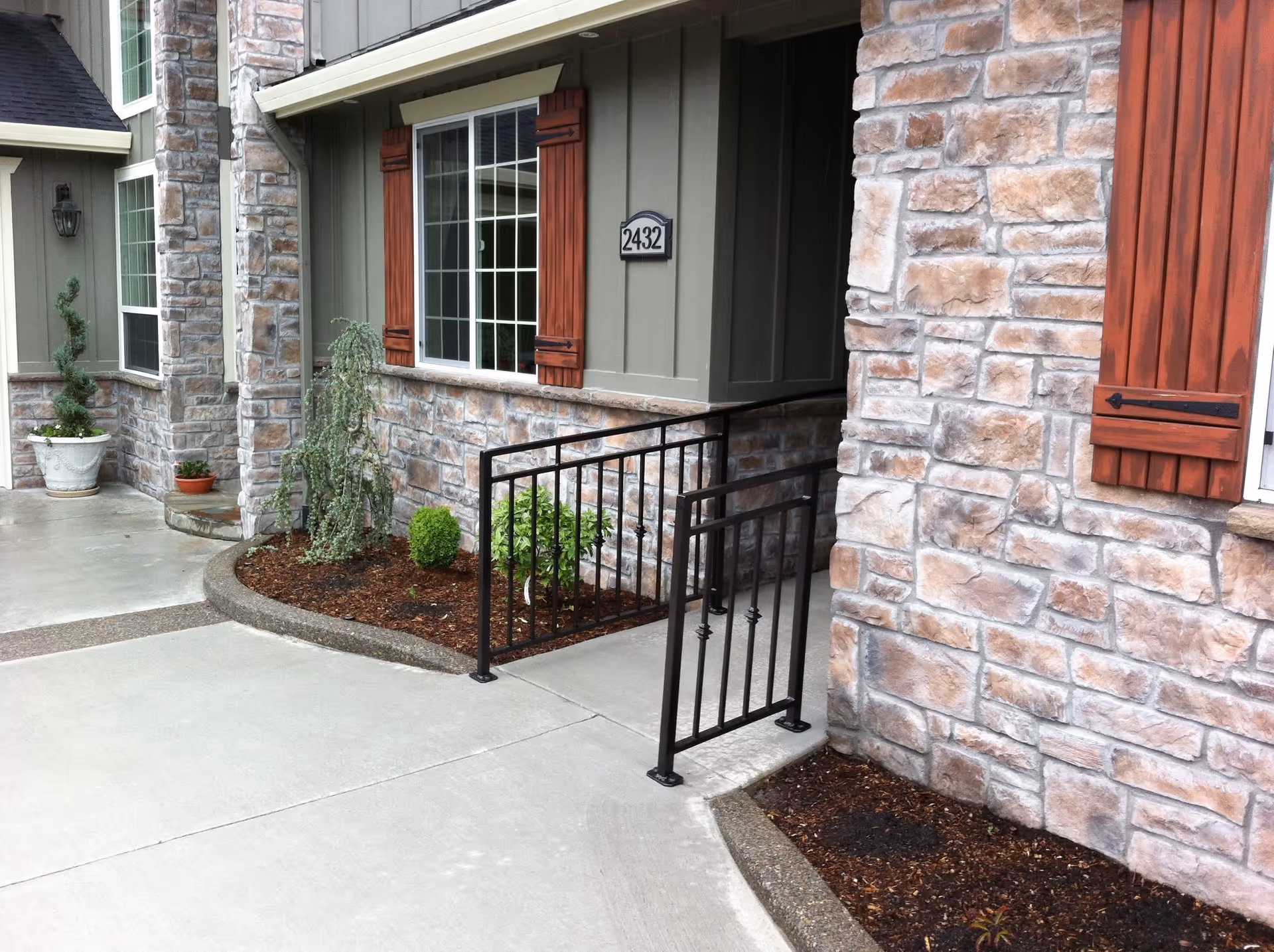 Exterior view of a building with stone and wood siding, a window with wooden shutters, a black metal handrail along a concrete walkway, and small landscaped areas with plants and mulch.