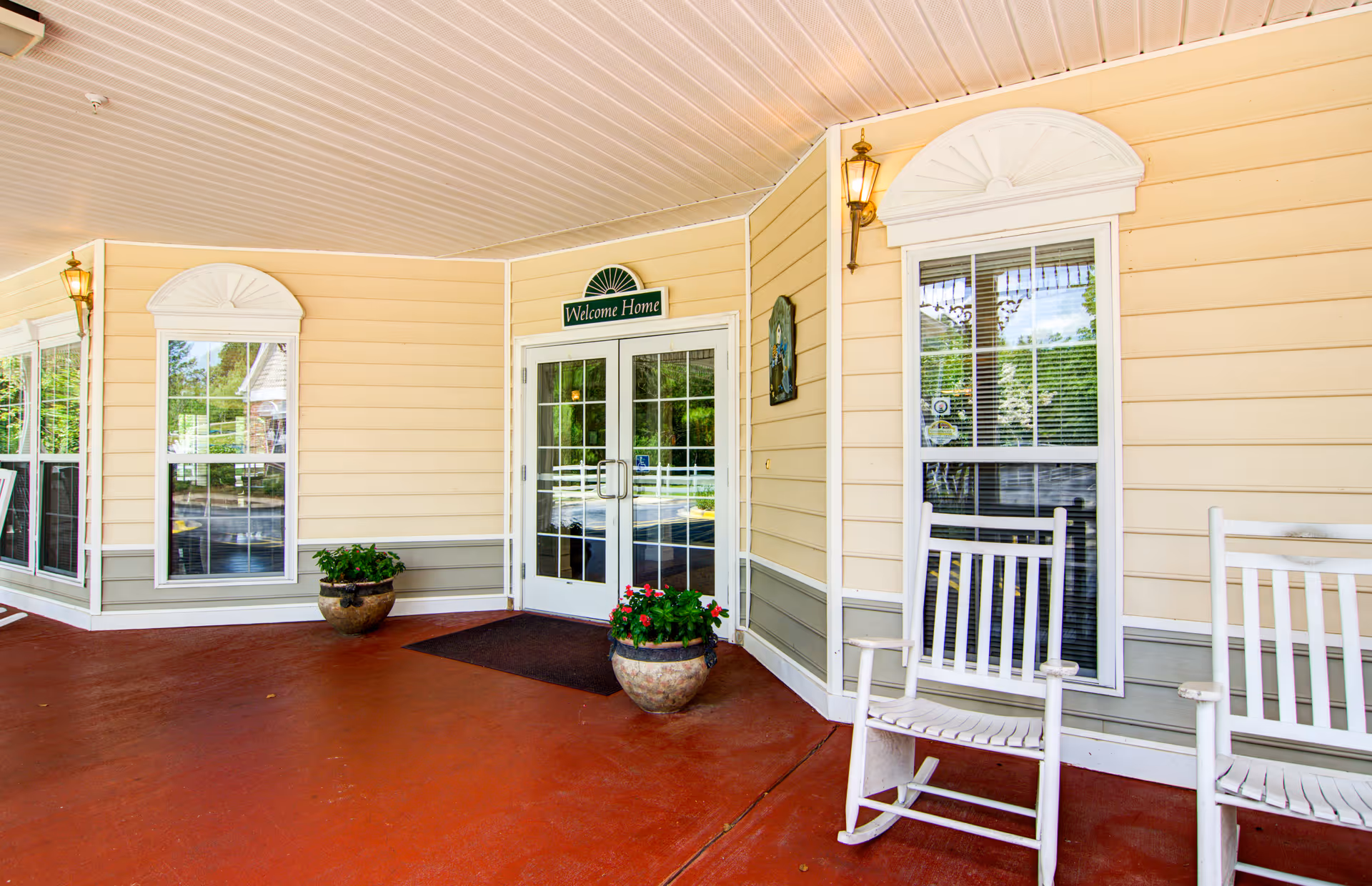 Covered entrance porch with double glass doors, a 'Welcome Home' sign, potted plants, and white rocking chairs.